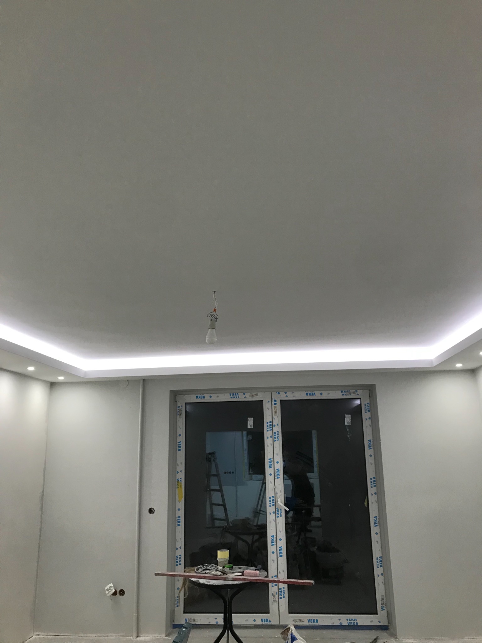 Pokój w trakcie remontu z zamontowanym sufitem podwieszanym z oświetleniem LED, widoczne narzędzia na stole i drabina w tle.