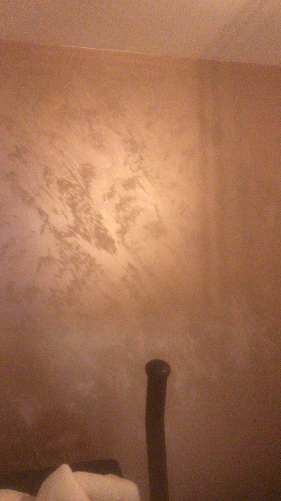 Ściana pokryta dekoracyjną, metaliczną farbą w odcieniach brązu i złota, z widoczną fakturą i delikatnymi smugami, na dole fragment czarnego przedmiotu przypominającego rurkę.