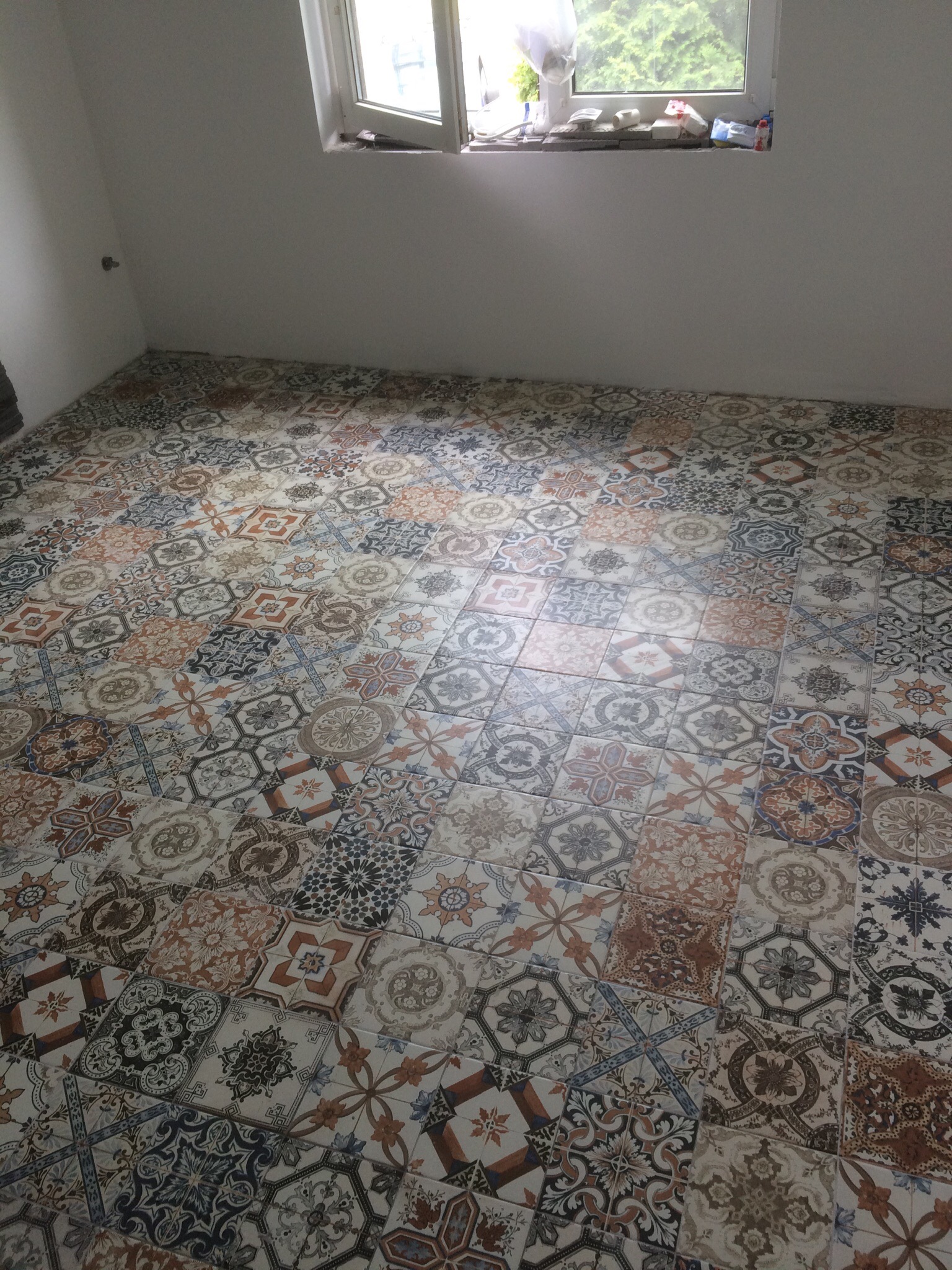 Ułożona podłoga z wzorzystych płytek ceramicznych w odcieniach brązu, beżu i szarości w pustym pomieszczeniu z oknem, na parapecie widoczne akcesoria budowlane.