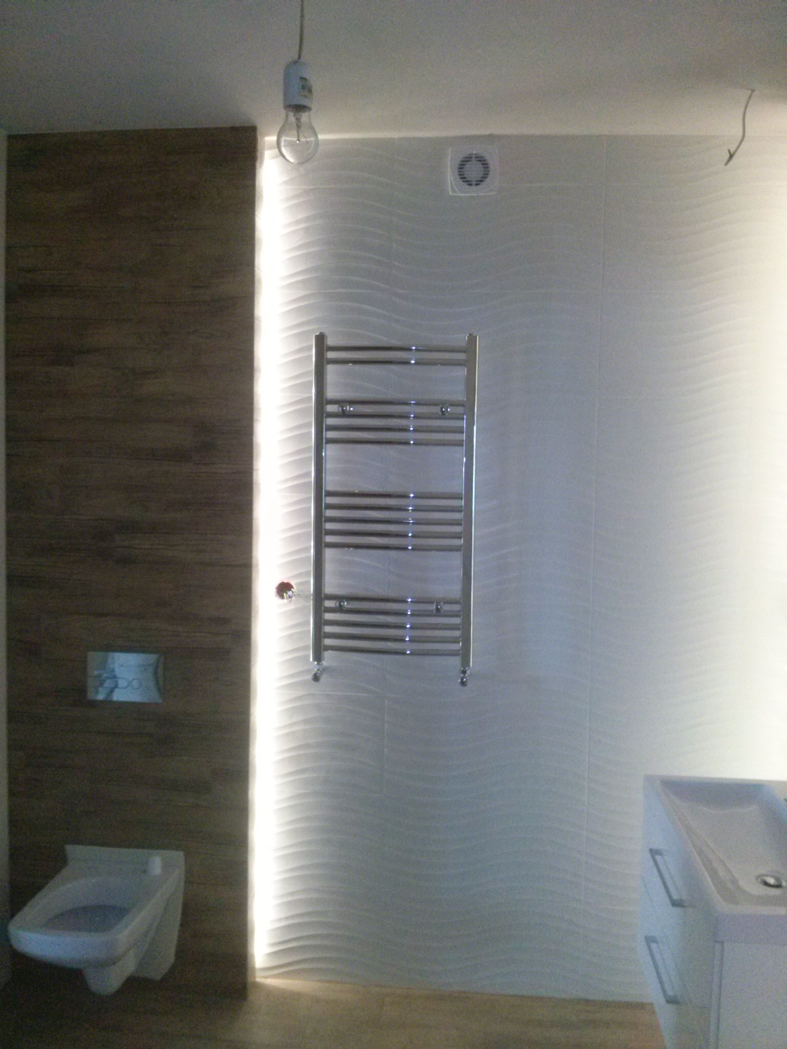 Nowoczesna łazienka z drewnopodobną okładziną ścienną, białe płytki 3D, grzejnik drabinkowy i podwieszana toaleta z przyciskiem spłukującym, oświetlona pionową listwą LED. Widoczna minimalistyczna...