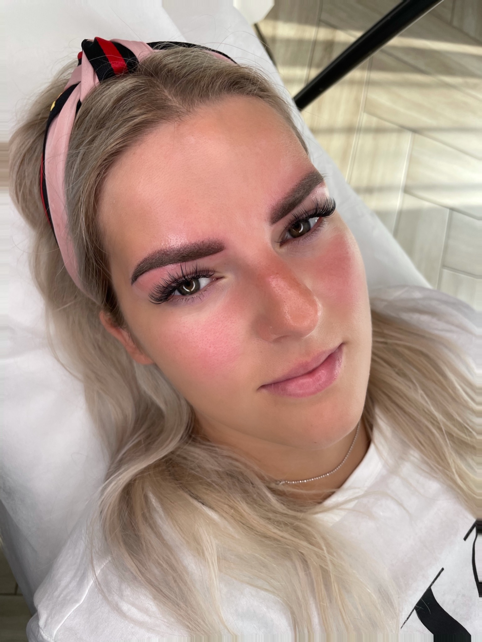 Młoda kobieta z blond włosami i różową opaską, leżąca na białym prześcieradle po zabiegu stylizacji brwi i przedłużania rzęs, z lekkim zaczerwienieniem skóry wokół brwi.