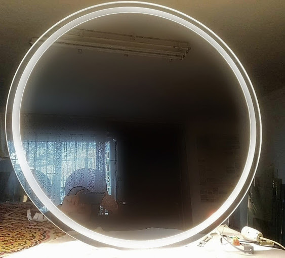 Okrągłe lustro z podświetleniem LED, leżące na stole. W odbiciu widać okno z zasłonami i osobę robiącą zdjęcie. Widoczne narzędzia szklarskie.