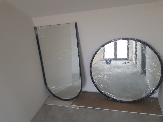 Dwa lustra: okrągłe i w kształcie łuku, zabezpieczone folią, oparte o ścianę w surowym pomieszczeniu. Widoczne odblaski okna.