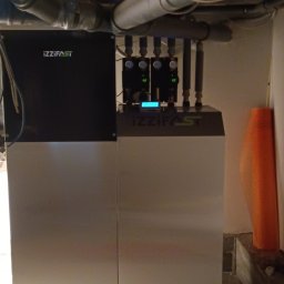 HYD-MAR - Nowoczesny piec grzewczy Izzifast z podłączonymi rurami i manometrami, zainstalowany w pomieszczeniu z widocznymi rurami pod sufitem i osłoną izolacyjną przy ścianie.