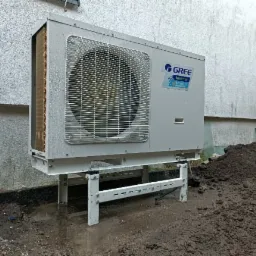 Zewnętrzna jednostka pompy ciepła Gree zamontowana na metalowej konstrukcji wsporczej przy ścianie budynku, widoczna świeżo wykopana ziemia.