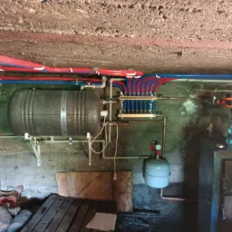 Instalacja grzewcza w piwnicy z zbiornikiem akumulacyjnym, rozdzielaczem, pompą obiegową i rurami w kolorach czerwonym i niebieskim pod betonowym sufitem. Widoczne elementy instalacji i narzędzia.