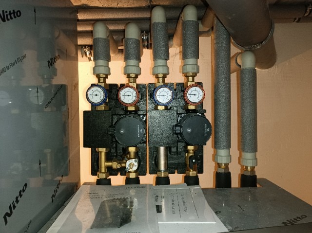 Rozdzielacz ogrzewania podłogowego z dwoma pompami obiegowymi, manometrami z czerwoną i niebieską obwódką, rurami w izolacji termicznej i widocznym zbiornikiem z folią ochronną.