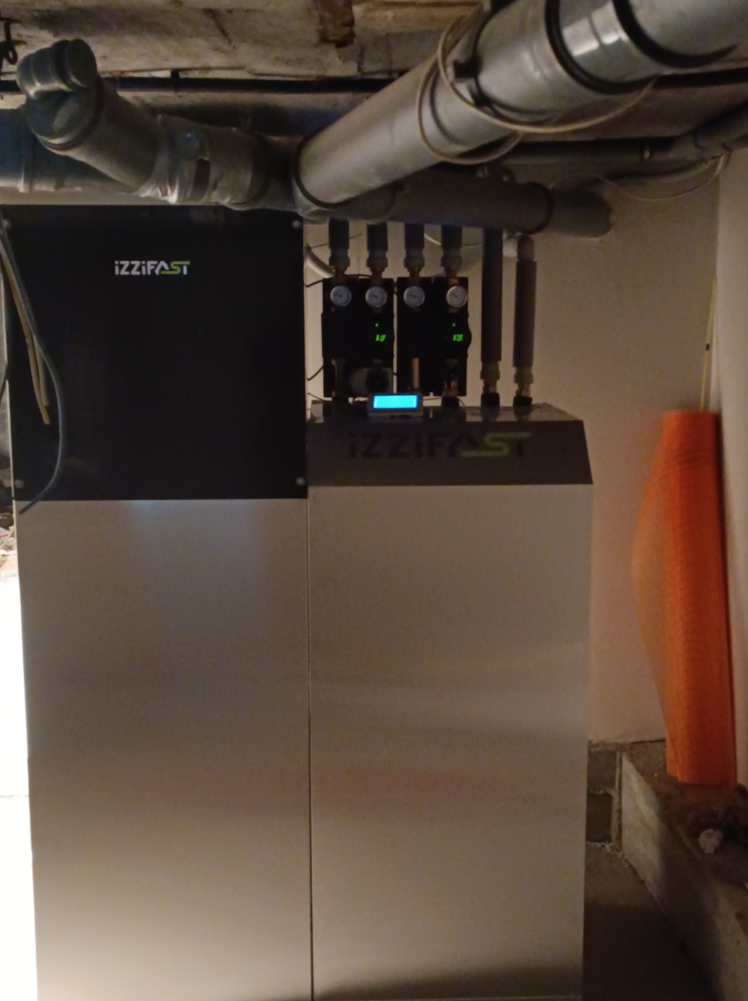 Nowoczesny piec grzewczy Izzifast z podłączonymi rurami i manometrami, zainstalowany w pomieszczeniu z widocznymi rurami pod sufitem i osłoną izolacyjną przy ścianie.