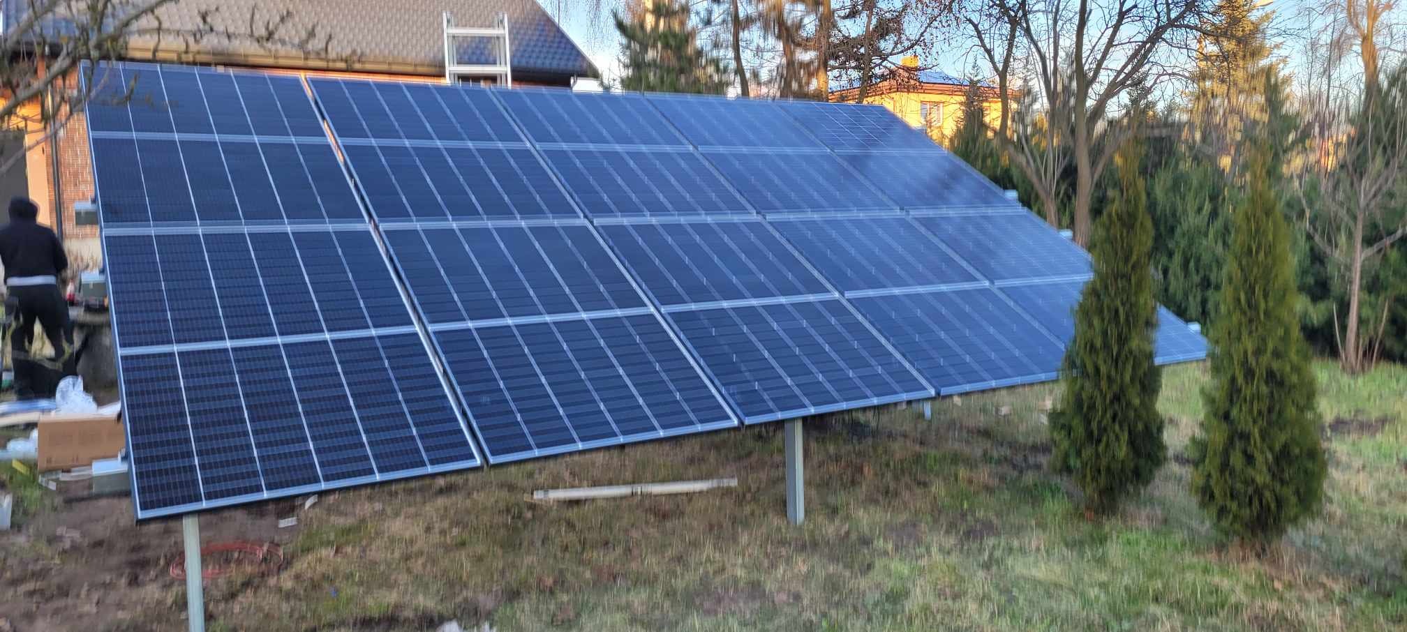 Montaż paneli fotowoltaicznych w ogrodzie, widoczny pracownik na drabinie przy panelach, w tle dom i drzewa.