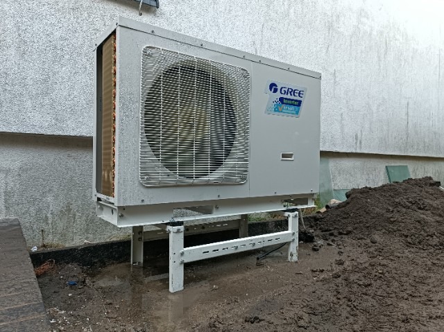 Zewnętrzna jednostka pompy ciepła Gree zamontowana na metalowej konstrukcji wsporczej przy ścianie budynku, widoczna świeżo wykopana ziemia.