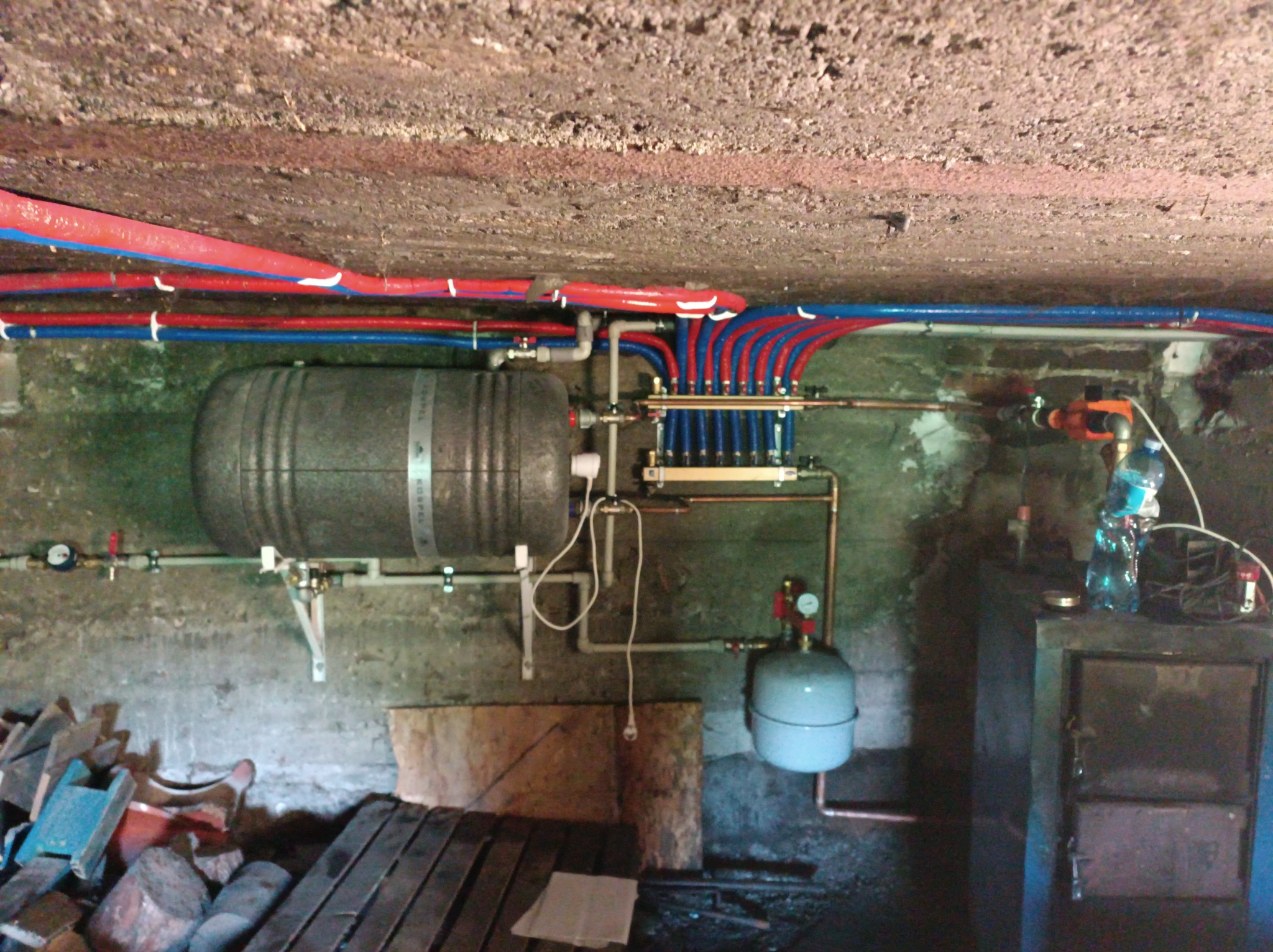 Instalacja grzewcza w piwnicy z zbiornikiem akumulacyjnym, rozdzielaczem, pompą obiegową i rurami w kolorach czerwonym i niebieskim pod betonowym sufitem. Widoczne elementy instalacji i narzędzia.