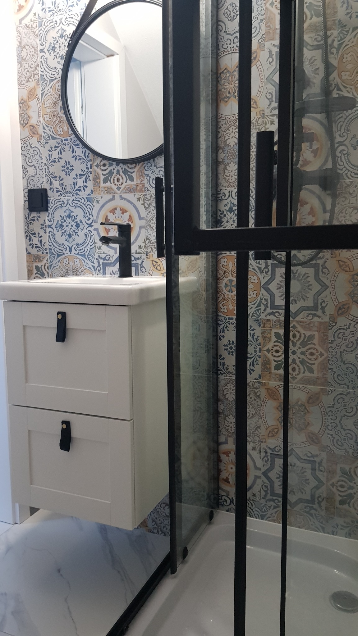 Nowoczesna łazienka z białą szafką pod umywalkę z czarnymi uchwytami, okrągłym lustrem w czarnej ramie, czarną baterią i kabiną prysznicową z czarnymi profilami na tle ściany wyłożonej wzorzystymi...