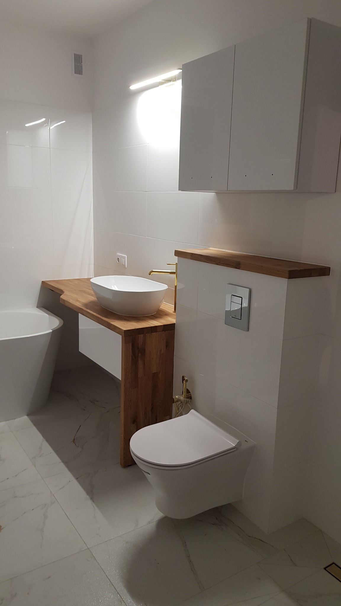 Nowoczesna łazienka z białą ceramiką, drewnianym blatem pod umywalką i zabudową WC ze srebrnym przyciskiem, białe płytki na ścianach i marmurowa podłoga.