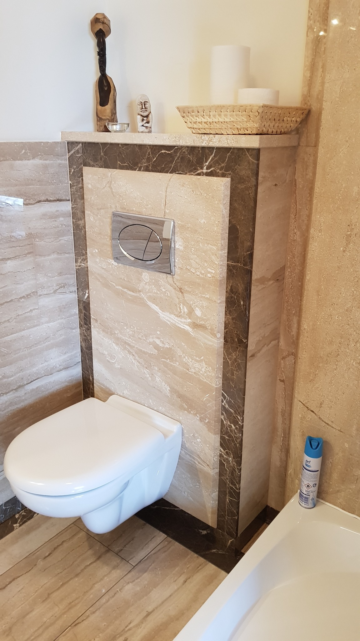 Zawieszona toaleta w łazience z beżowymi płytkami imitującymi marmur, z dekoracyjną obudową stelaża i półką z ozdobami: drewniana figurka, kamienna rzeźba i koszyk na papier toaletowy.