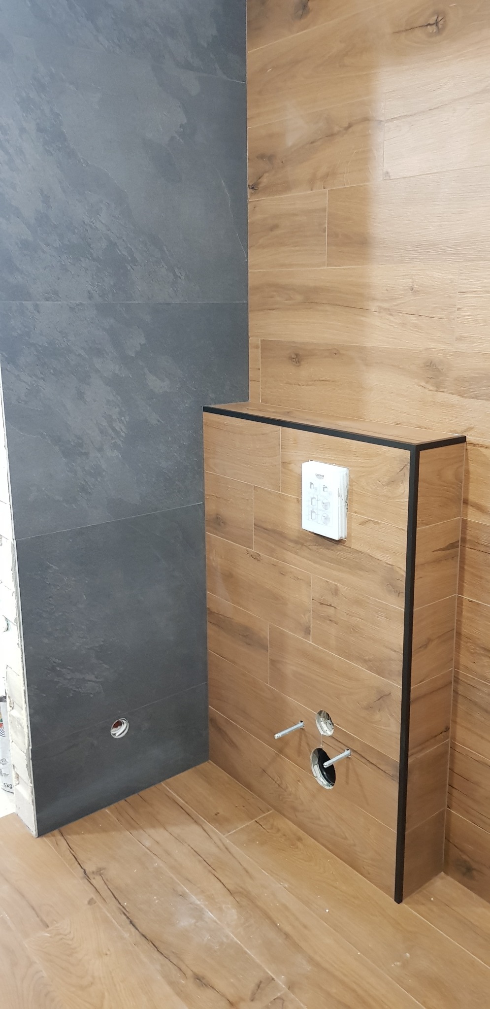 Ściana wyłożona szarymi płytkami imitującymi kamień i zabudowa stelaża WC wykończona płytkami drewnopodobnymi w trakcie montażu, widoczne wystające rurki i przycisk spłukujący Grohe.