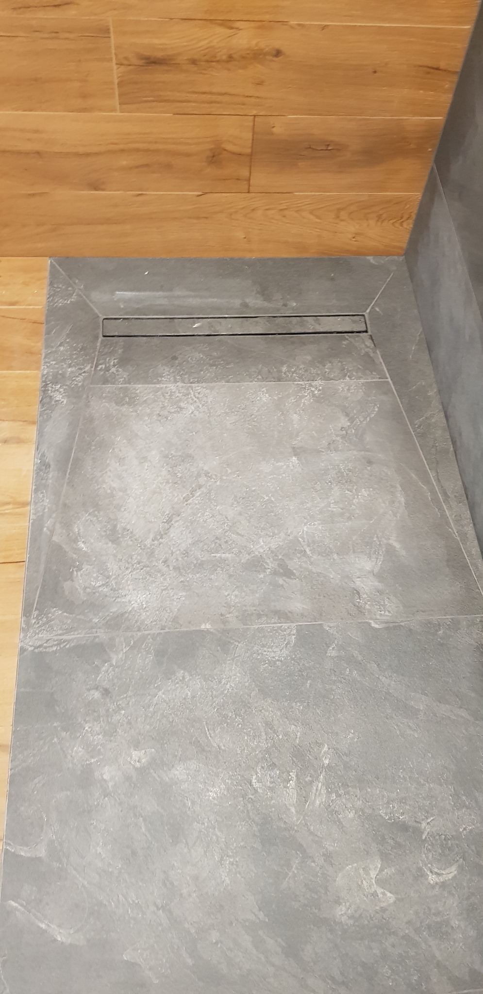 Szary brodzik z odpływem liniowym, ułożony na podłodze z paneli drewnopodobnych, widok z góry.