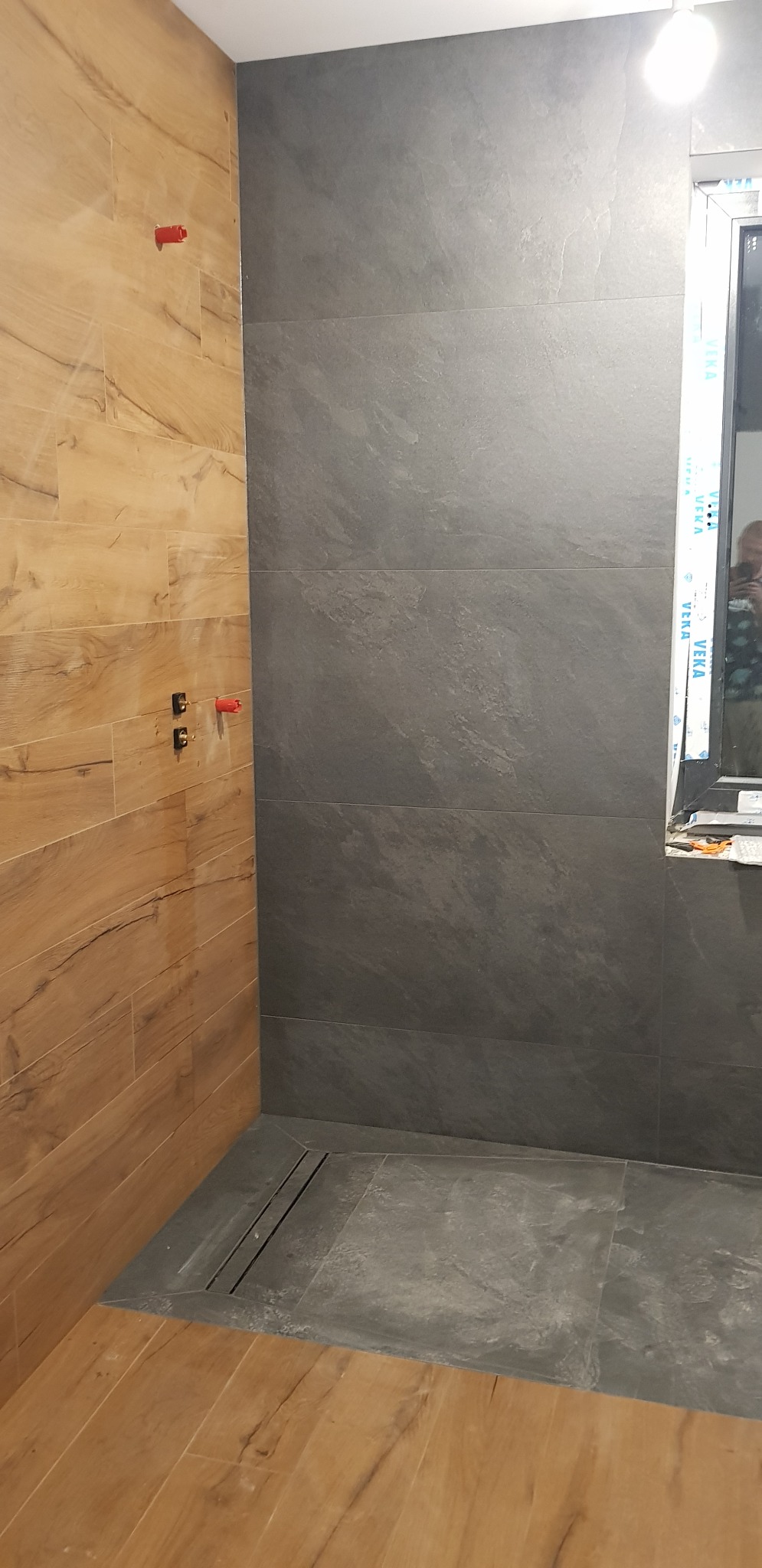 Wykończona kabina prysznicowa z odpływem liniowym, wyłożona płytkami imitującymi drewno i szarym kamieniem, widoczne elementy instalacji wodnej.