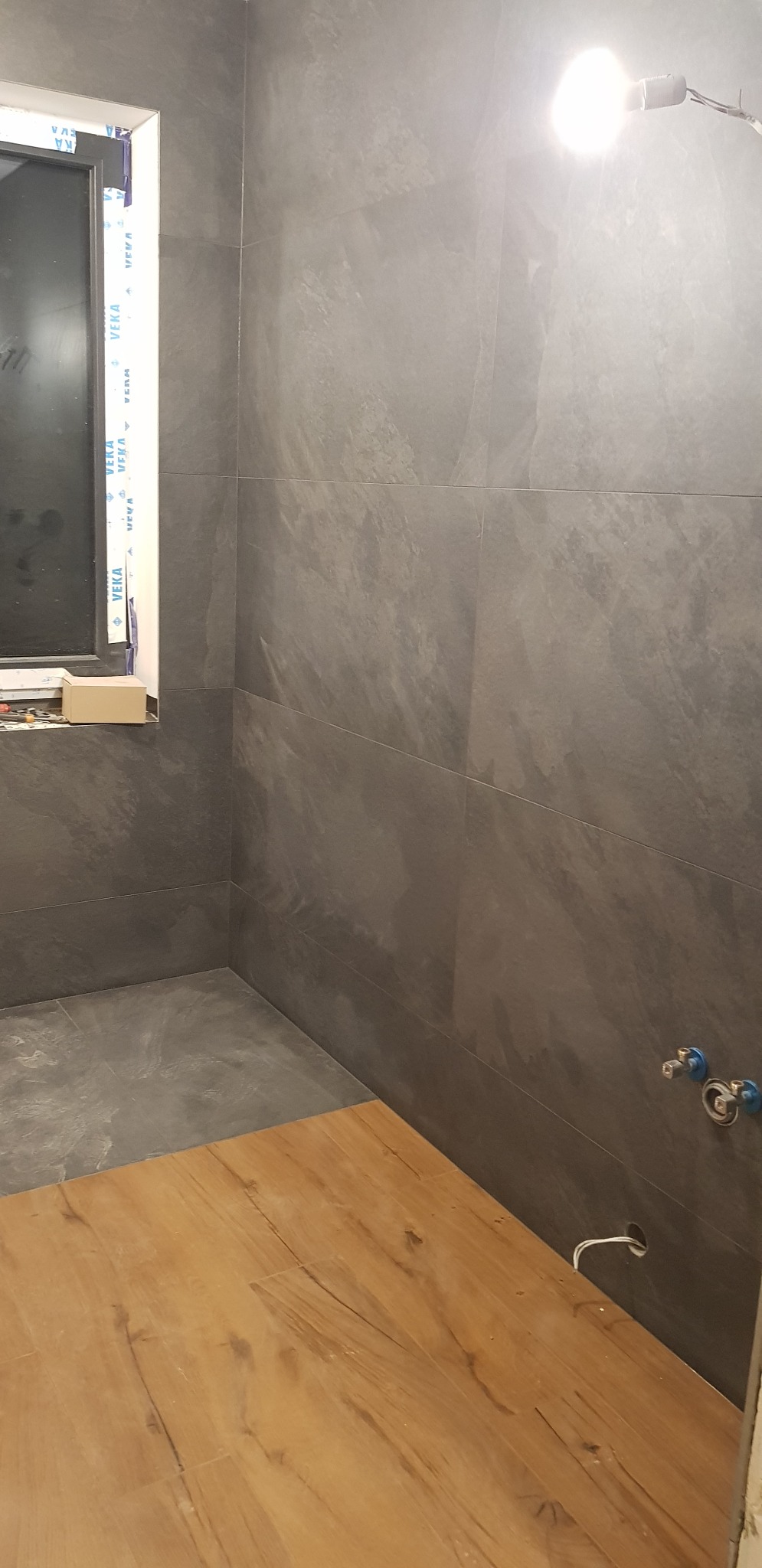 Szara łazienka w trakcie wykończenia: ściany wyłożone płytkami imitującymi kamień, częściowo ułożona podłoga z paneli drewnopodobnych, widoczne przyłącza wodne i okno z folią ochronną.