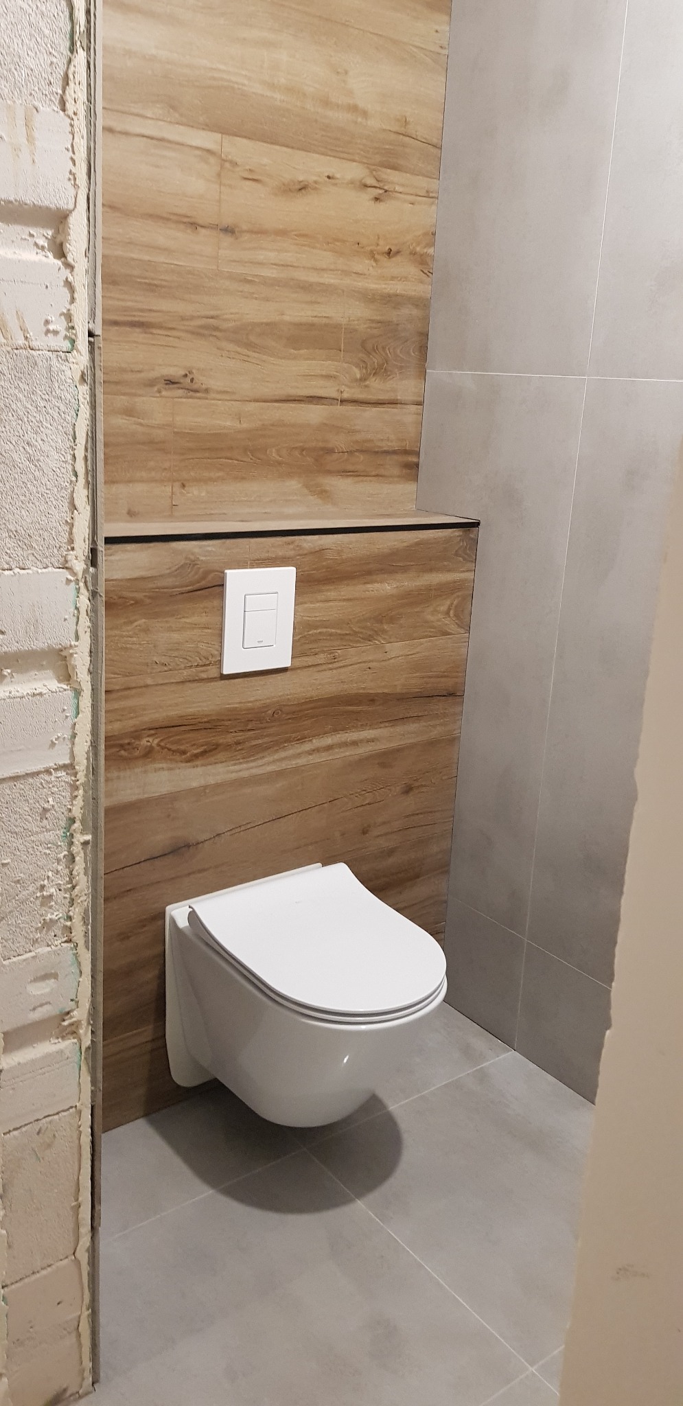 Wisząca toaleta z deską wolnoopadającą zamontowana na ścianie wyłożonej płytkami imitującymi drewno, obok ściana z szarych płytek i surowy fragment ściany w trakcie remontu.