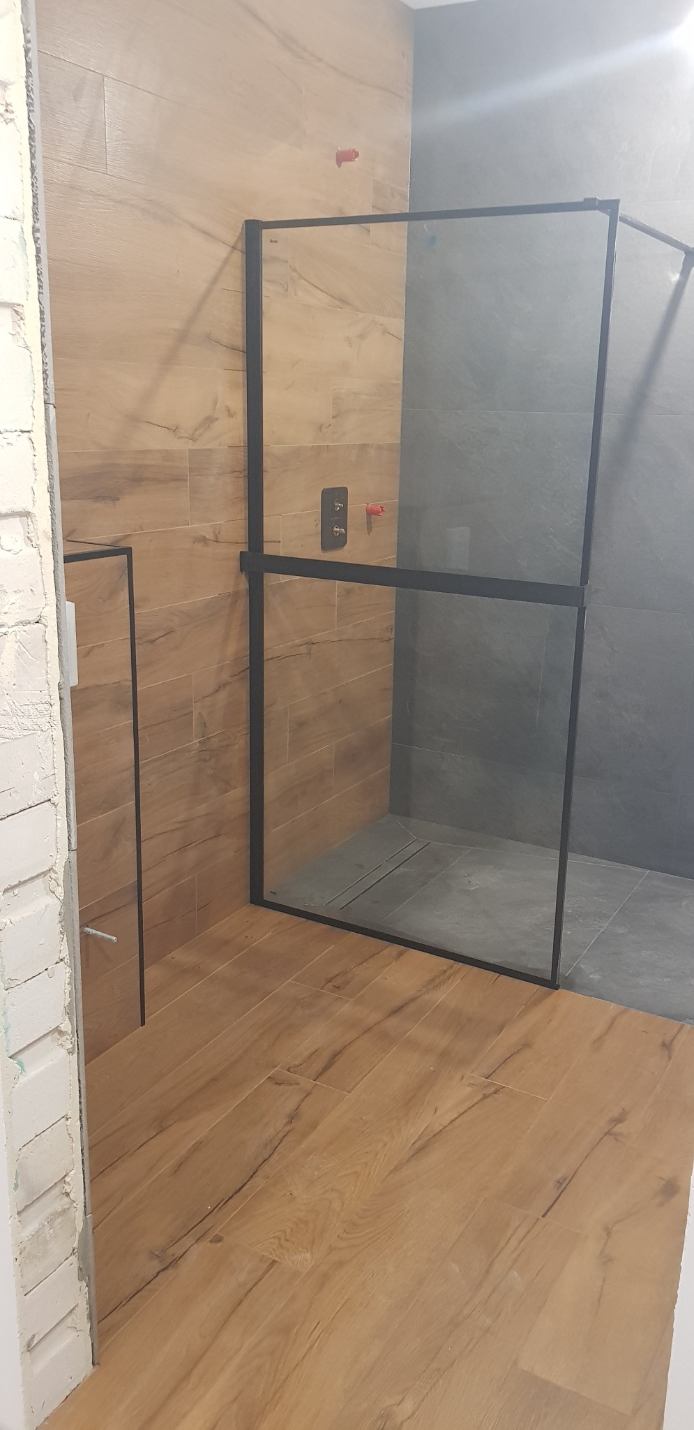 Częściowo wykończona łazienka z kabiną prysznicową z czarną ramą, drewnopodobne płytki na ścianie i podłodze, szare płytki wewnątrz kabiny, widoczne elementy instalacji.