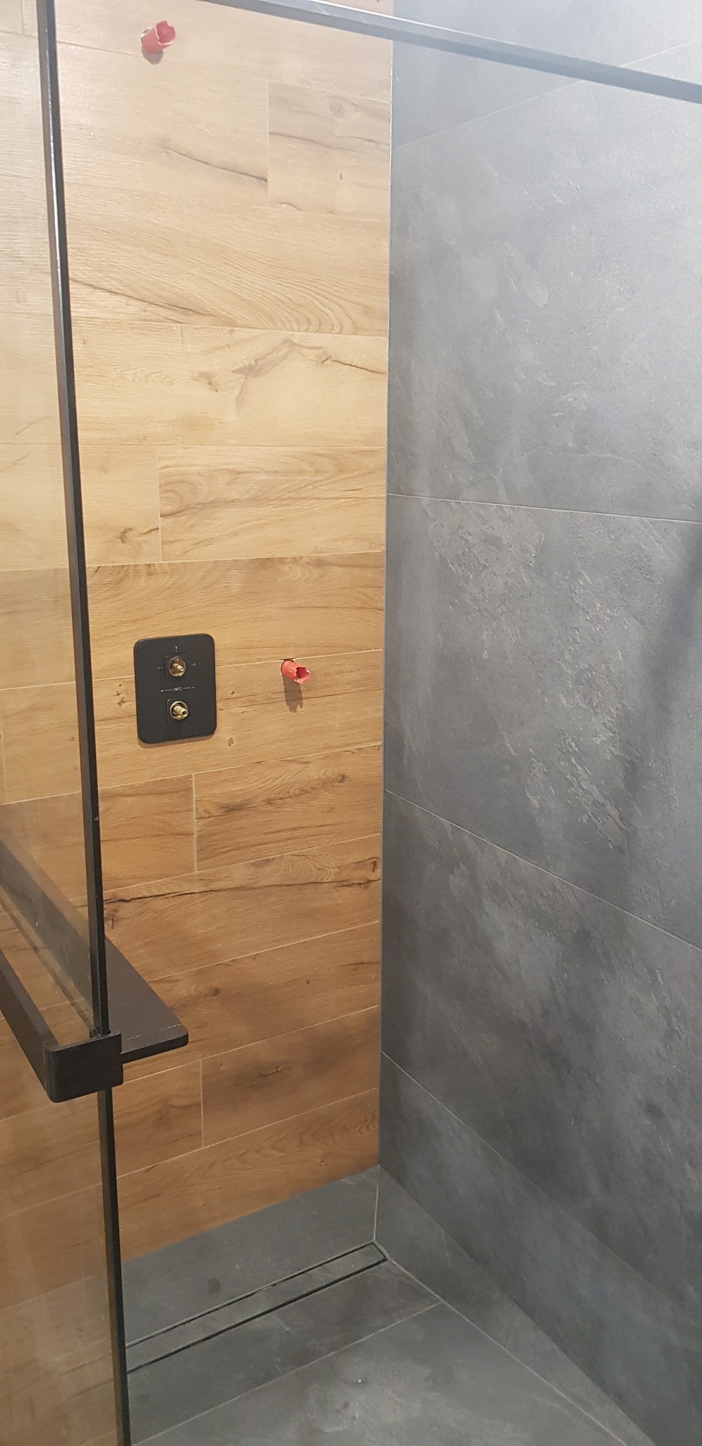Wykończona kabina prysznicowa z drewnopodobnymi płytkami na ścianie z instalacją podtynkową i szarą płytką imitującą kamień, widoczny odpływ liniowy.