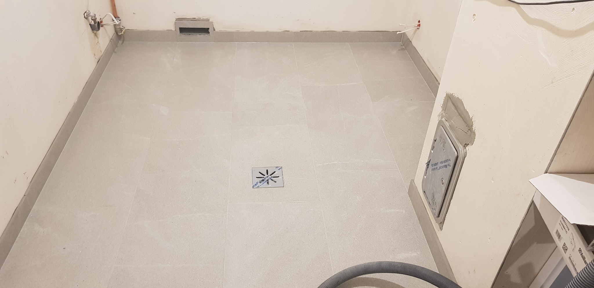 Wykończona podłoga w łazience z jasnych, kwadratowych płytek, centralnie umieszczony odpływ liniowy z metalową kratką, widoczne przyłącza wodne i wentylacyjne, oraz szary wąż odkurzacza.