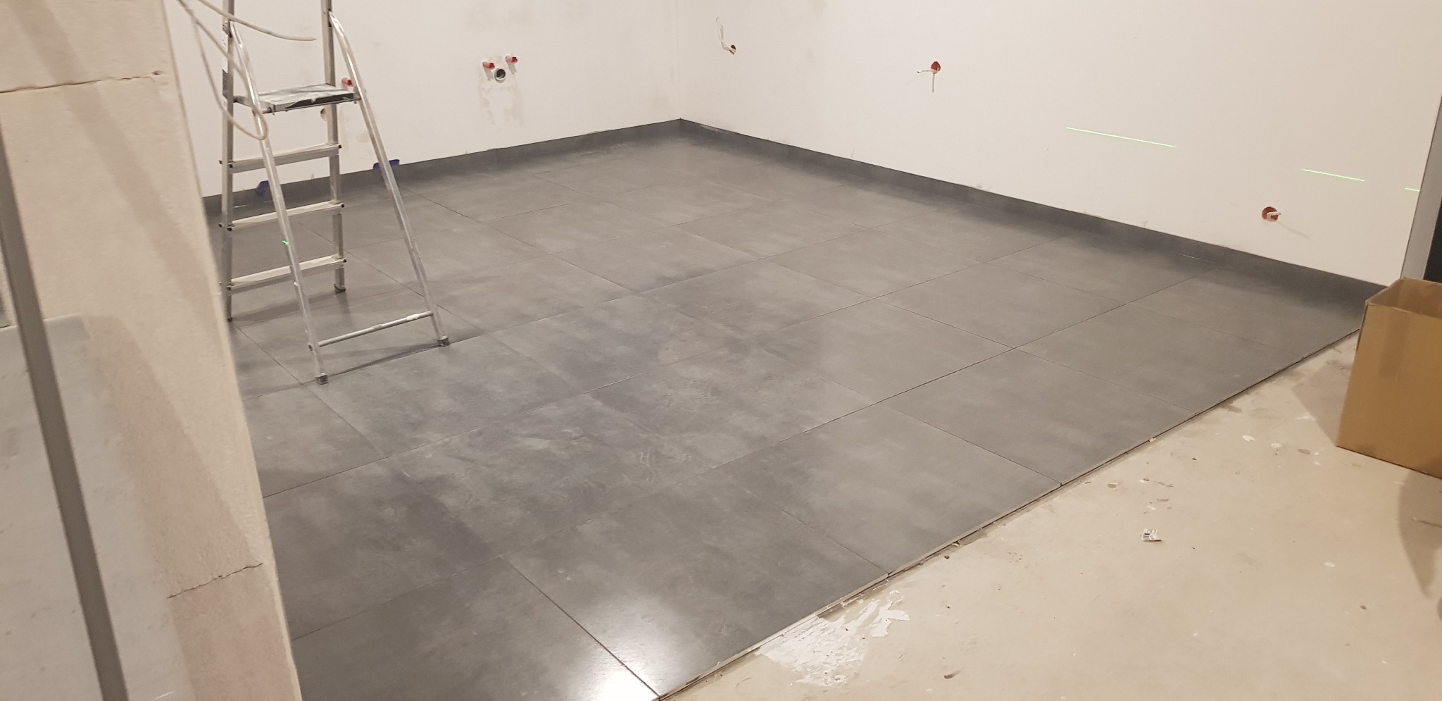 Ułożone szare płytki podłogowe w surowym wnętrzu, widoczna drabina, laserowy poziom i wystające kable ze ściany.
