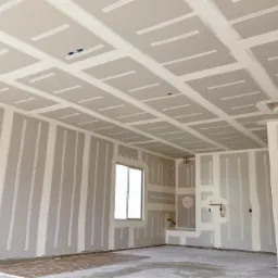 Wnętrze w trakcie prac: ściany i sufit pokryte płytami gipsowo-kartonowymi, łączenia wzmocnione taśmą, widoczne puszki elektryczne i instalacja wodna, surowa podłoga betonowa.