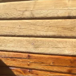 Fragment drewnianej ściany z bali, część pomalowana na jasny kolor, a dolna część w odcieniach brązu, widoczne słoje drewna i cień.