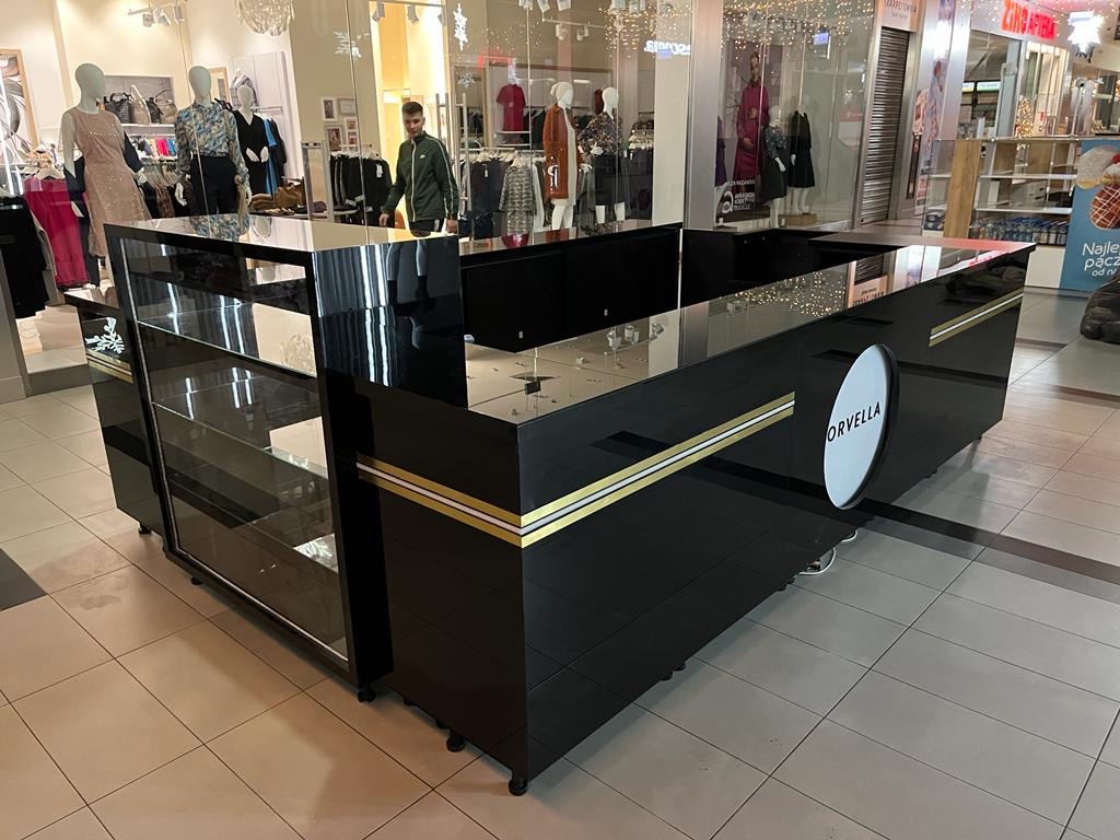 Czarna wyspa handlowa z logo Orvella w centrum handlowym, z przeszkloną witryną i złotymi akcentami. Widoczny fragment chodnika i manekiny w tle.
