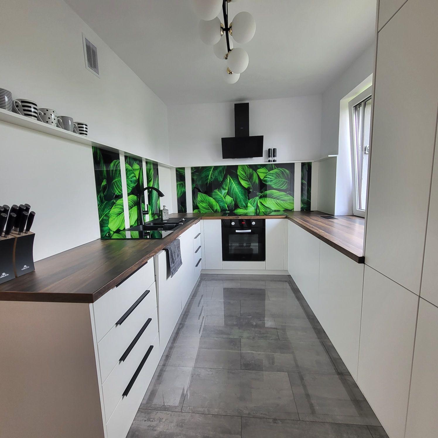 Nowoczesna kuchnia w bieli z drewnianym blatem i zielonym panelem ściennym z motywem liści. Minimalistyczne uchwyty szafek, szara podłoga, oświetlenie sufitowe.