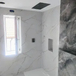 Wykończona łazienka z płytkami imitującymi marmur, otwartym oknem i czarną deszczownicą sufitową. Widoczna wnęka w ścianie i biały brodzik.