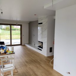 Wykonawstwo robót budowlanych - Salon po remoncie w Ciechanowie: panele podłogowe, marmurowy kominek, świeżo pomalowane ściany i widok na ogród przez duże okno.