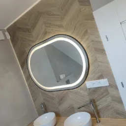 Nowoczesna łazienka po remoncie: umywalki nablatowe, lustro z podświetleniem LED i płytki ułożone w jodełkę. Widoczny fragment blatu i gniazdko elektryczne.