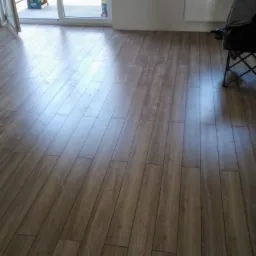 Ułożone panele podłogowe w odcieniach brązu, widoczne łączenia, z fragmentem balkonu w tle i krzesłem turystycznym w rogu pokoju.