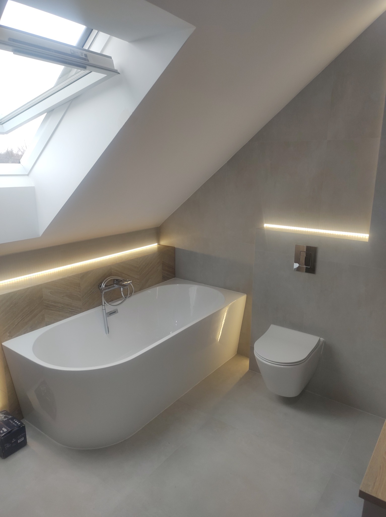 Nowoczesna łazienka na poddaszu z białą wolnostojącą wanną, podwieszaną toaletą i subtelnym oświetleniem LED pod skosem dachu i nad stelażem podtynkowym.