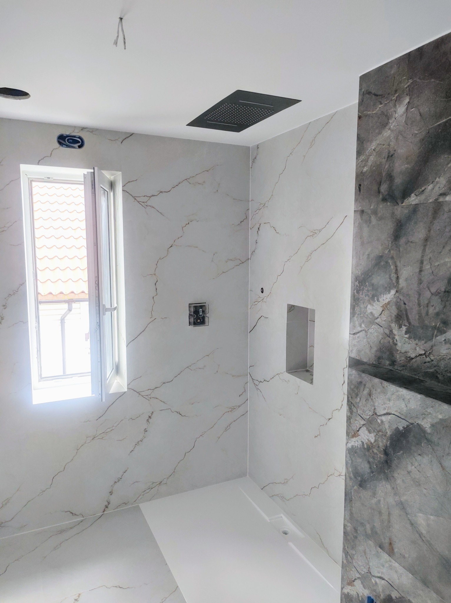 Wykończona łazienka z płytkami imitującymi marmur, otwartym oknem i czarną deszczownicą sufitową. Widoczna wnęka w ścianie i biały brodzik.