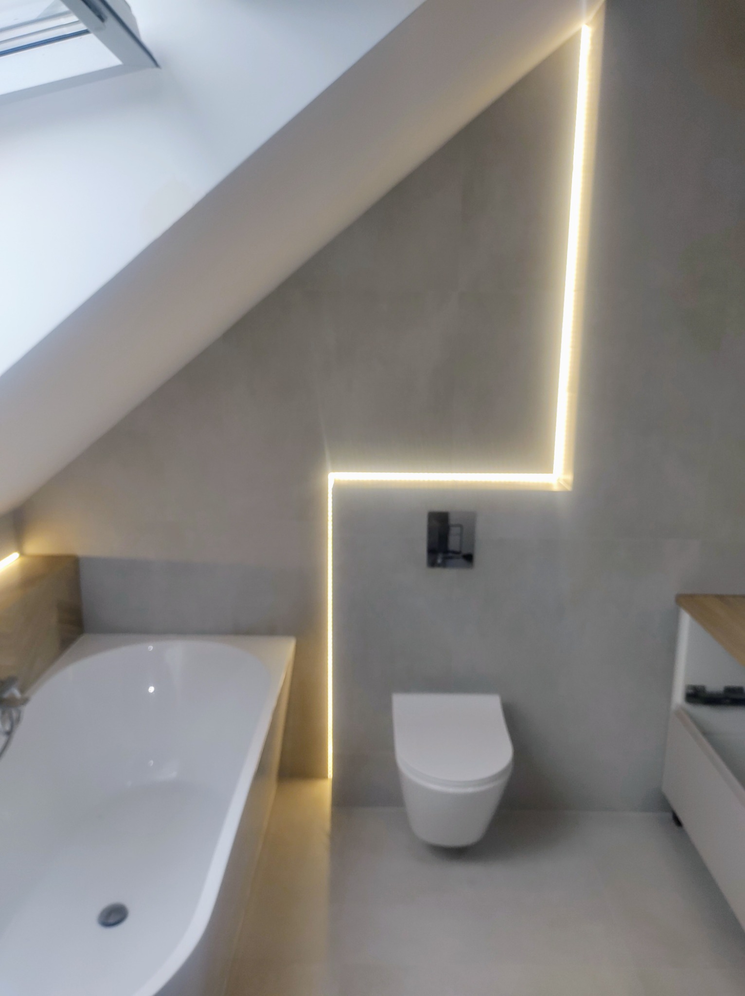 Jasna łazienka na poddaszu z wanną, toaletą podwieszaną i dekoracyjnym oświetleniem LED wzdłuż ścian.