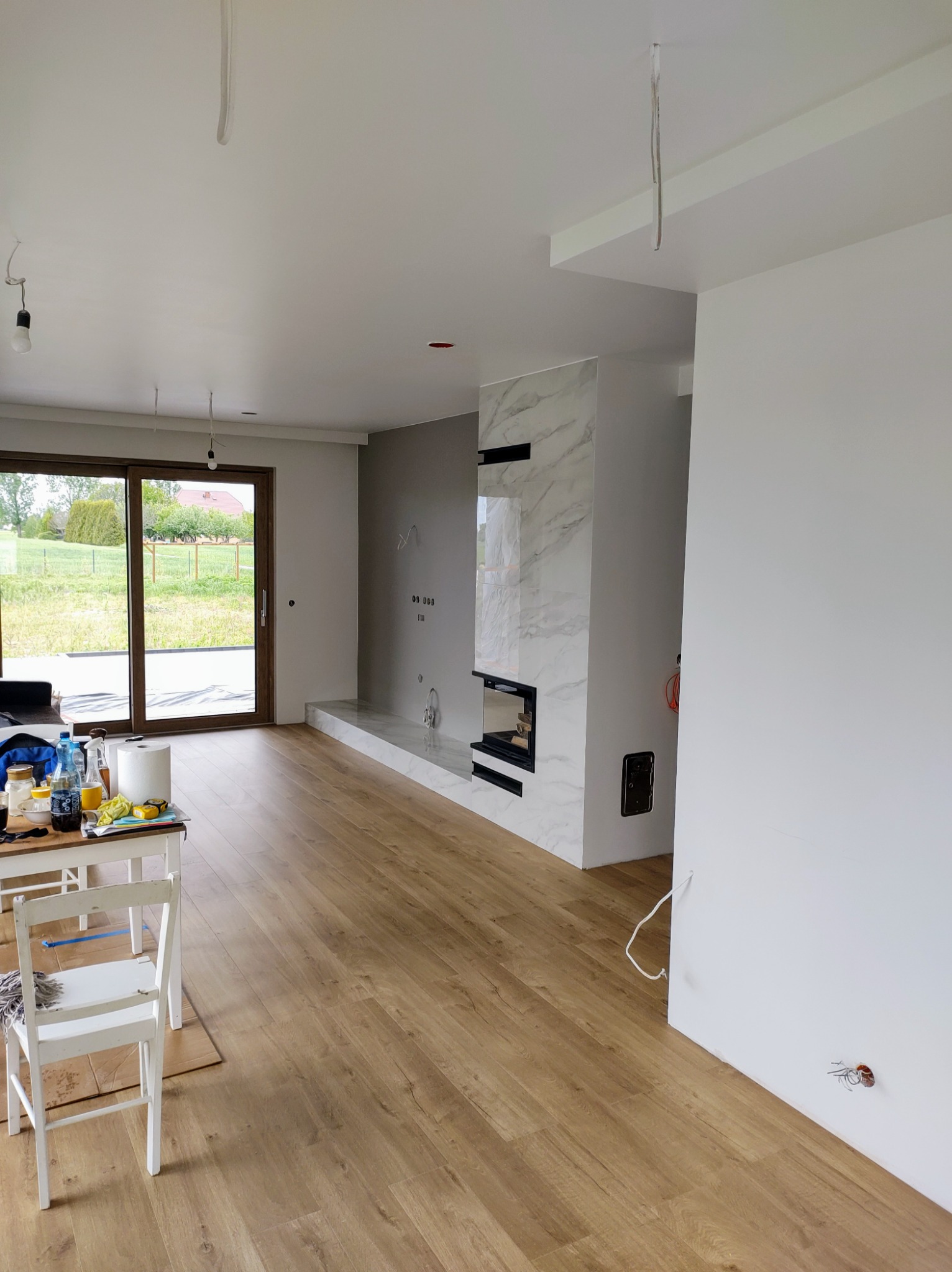 Salon po remoncie w Ciechanowie: panele podłogowe, marmurowy kominek, świeżo pomalowane ściany i widok na ogród przez duże okno.