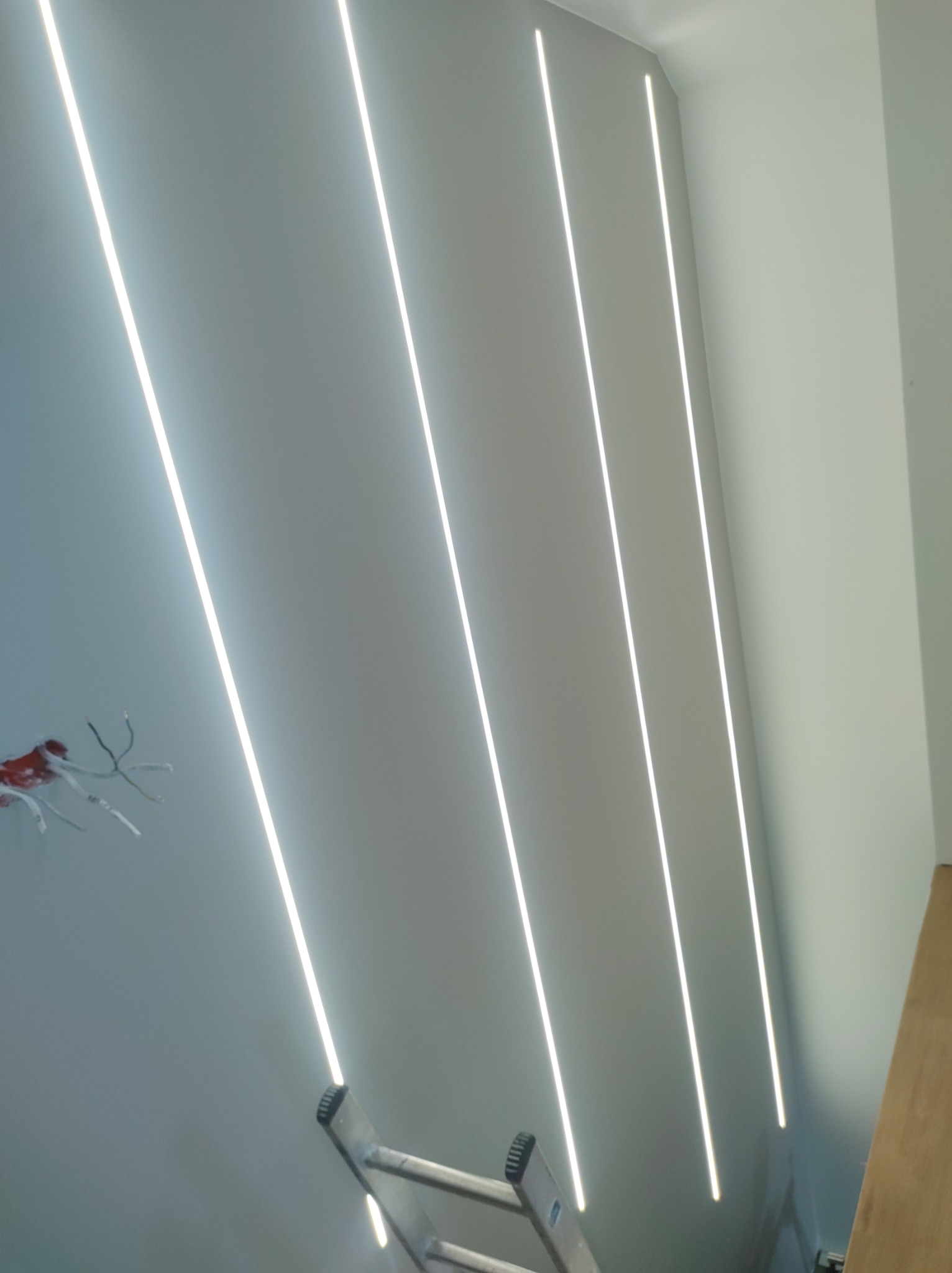 Nowoczesne oświetlenie sufitowe z liniowymi lampami LED w minimalistycznym wnętrzu. Widoczne przewody i drabina, sugerujące etap instalacji.
