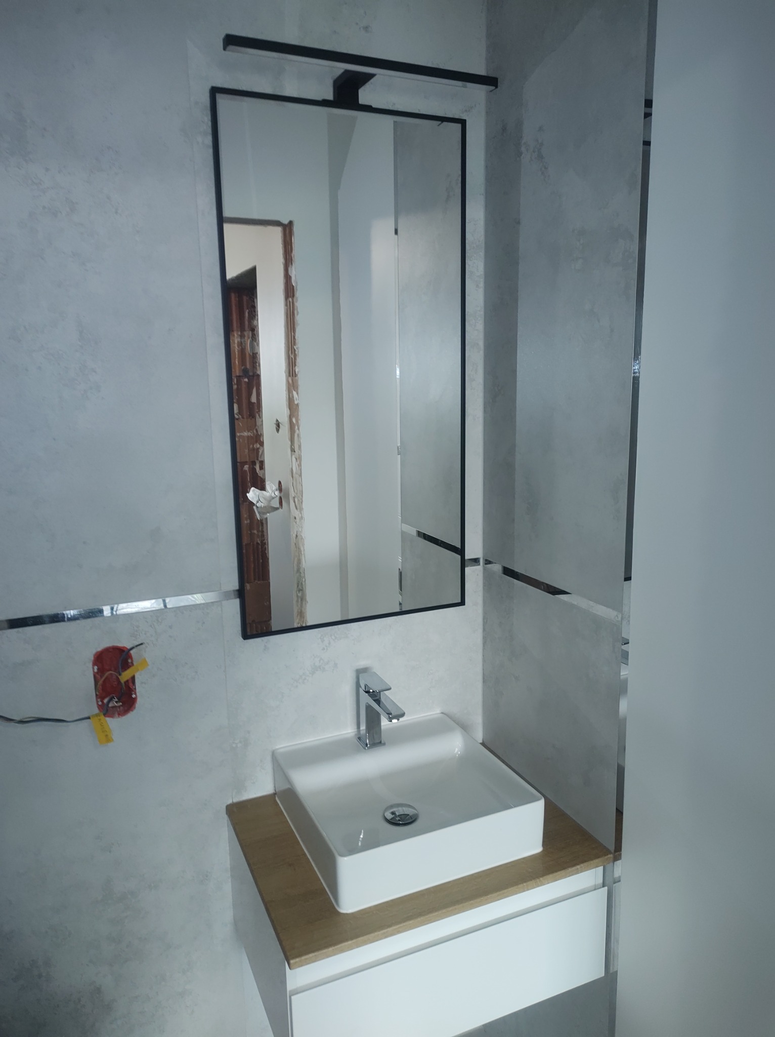 Nowoczesna łazienka po remoncie: biała umywalka na drewnianym blacie, duże lustro w czarnej ramie z oświetleniem. Widoczna surowa ściana w odbiciu.