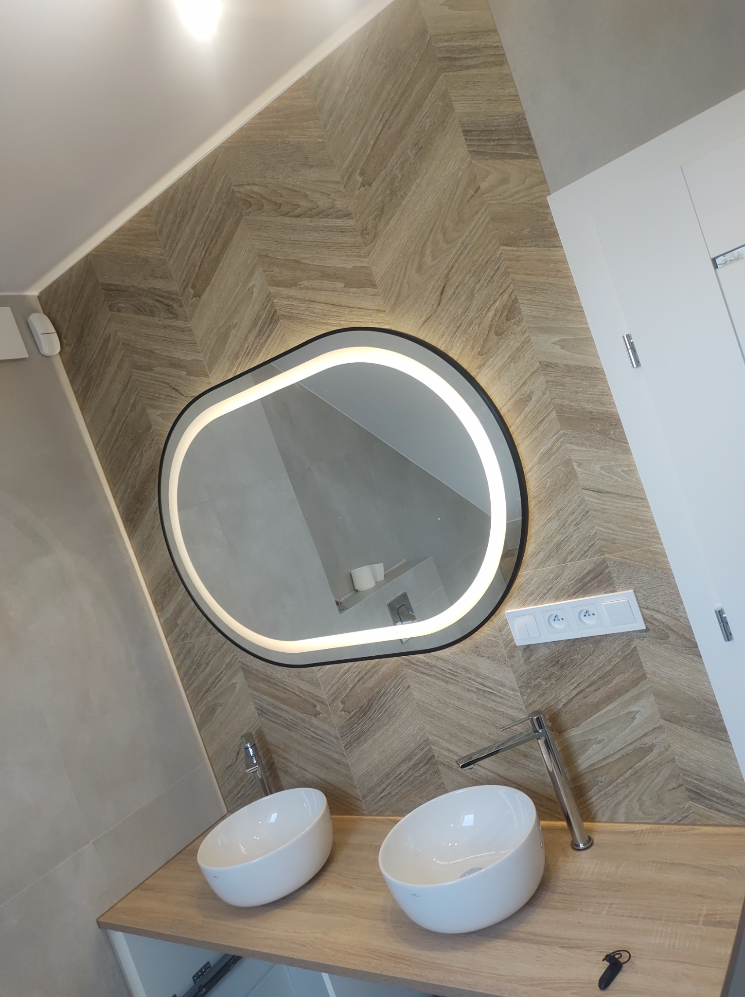 Nowoczesna łazienka po remoncie: umywalki nablatowe, lustro z podświetleniem LED i płytki ułożone w jodełkę. Widoczny fragment blatu i gniazdko elektryczne.