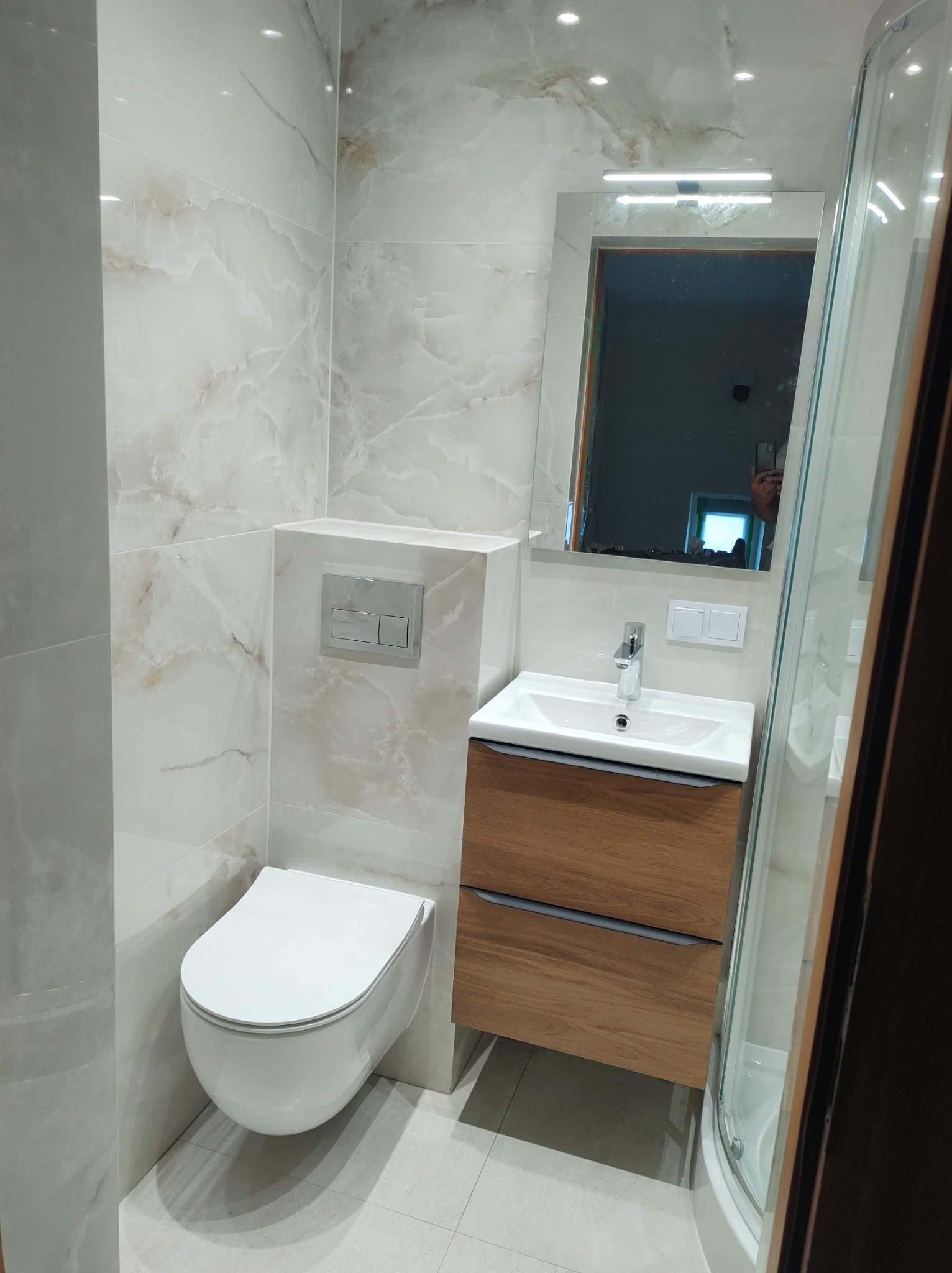 Nowoczesna łazienka z umywalką w drewnianej szafce, podwieszaną toaletą i kabiną prysznicową, wykończona płytkami imitującymi marmur.