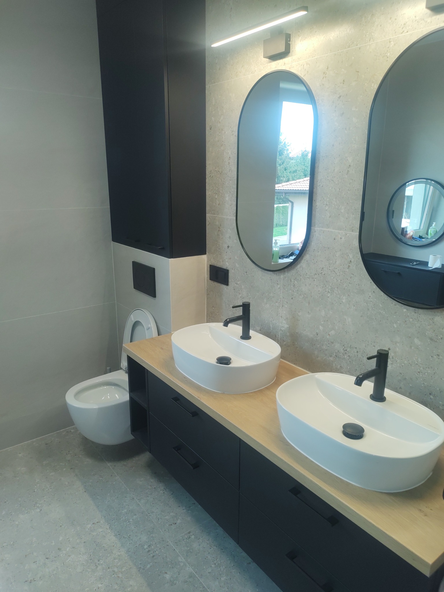 Nowoczesna łazienka z dwoma owalnymi umywalkami na drewnianym blacie, czarne szafki, lustra w czarnych ramach i szare płytki na ścianach i podłodze.