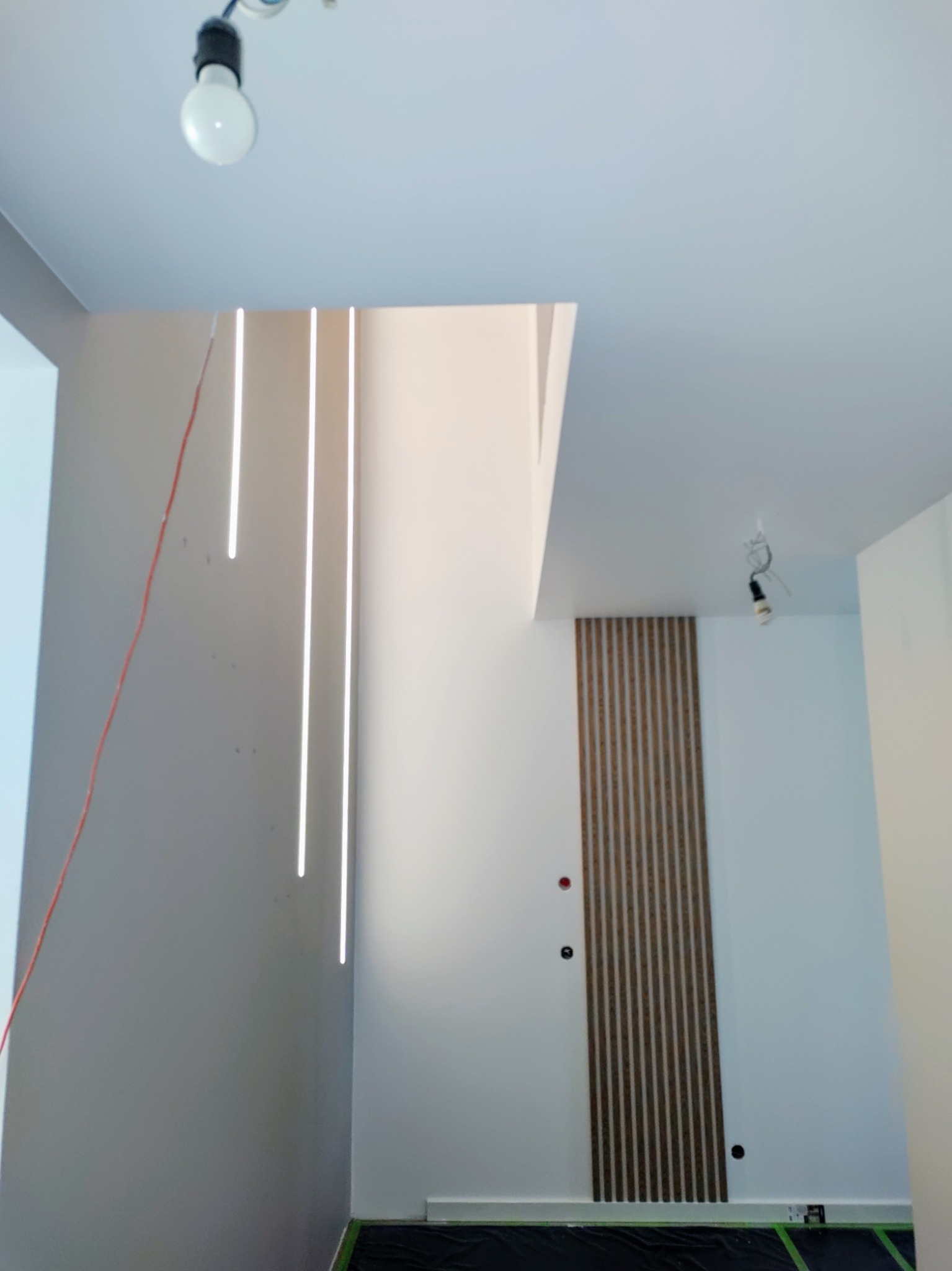 Nowoczesne oświetlenie LED w pionowych liniach na szarej ścianie, uzupełnione drewnianą dekoracją ścienną. Widoczne kable i tymczasowe oświetlenie sufitowe.