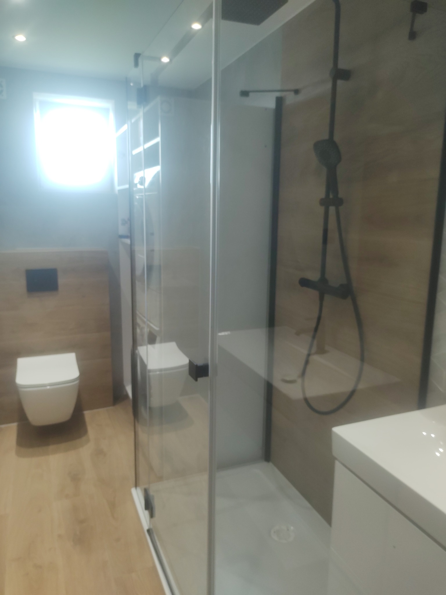 Nowoczesna łazienka z kabiną prysznicową typu walk-in, drewnopodobnymi płytkami na ścianach i podłodze oraz białymi sanitariatami. Minimalistyczny design, czarne akcenty.