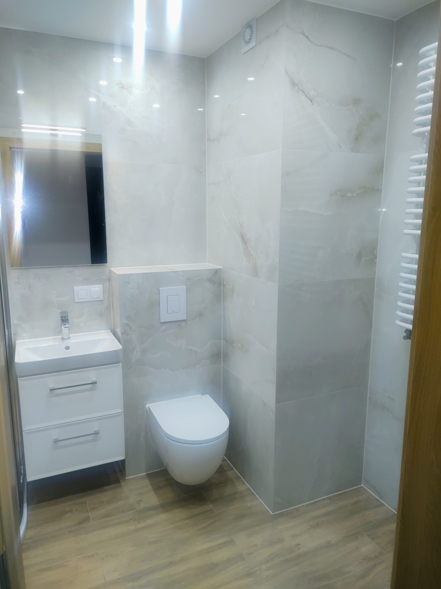 Nowoczesna łazienka z białymi płytkami imitującymi marmur, podwieszaną toaletą i drewnopodobną podłogą. Umywalka z szafką i lustrem, grzejnik drabinkowy.