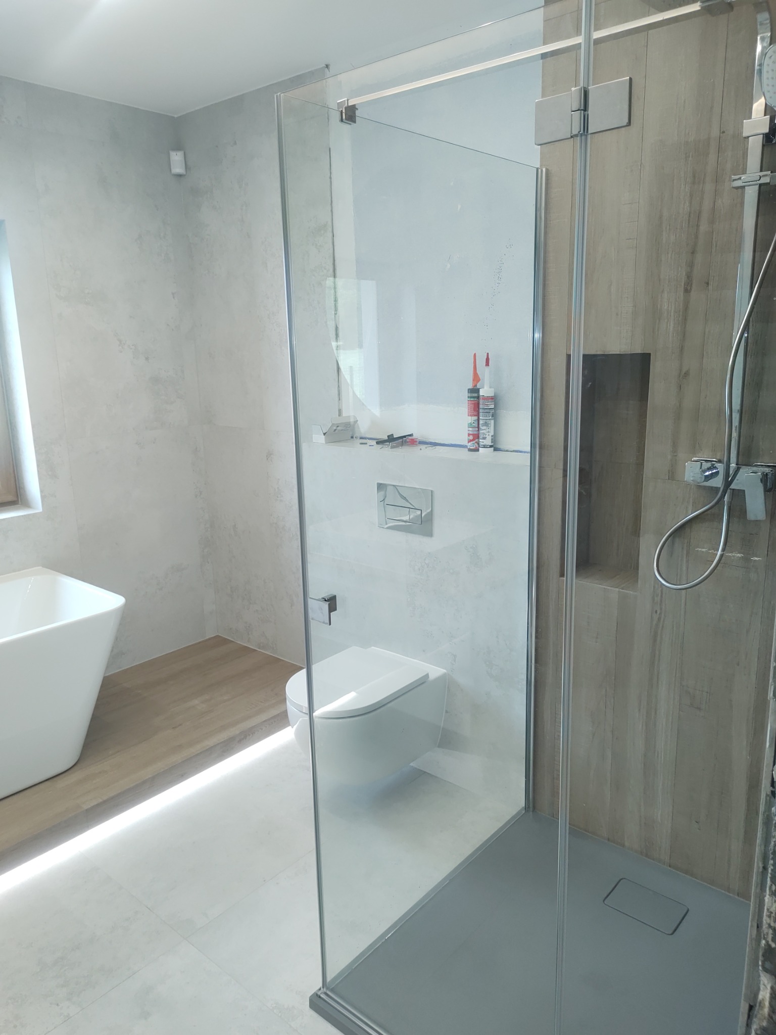 Nowoczesna łazienka z kabiną prysznicową typu walk-in, białą armaturą i płytkami imitującymi beton. Widoczne detale wykończeniowe i oświetlenie LED.