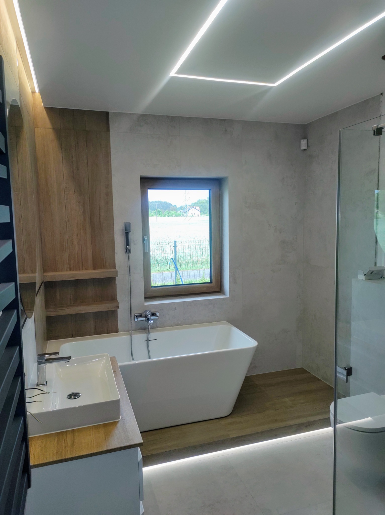 Nowoczesna łazienka z wolnostojącą wanną, szklaną kabiną prysznicową i drewnianymi akcentami. Okno z widokiem na zieleń. Oświetlenie LED podkreśla minimalistyczny design.