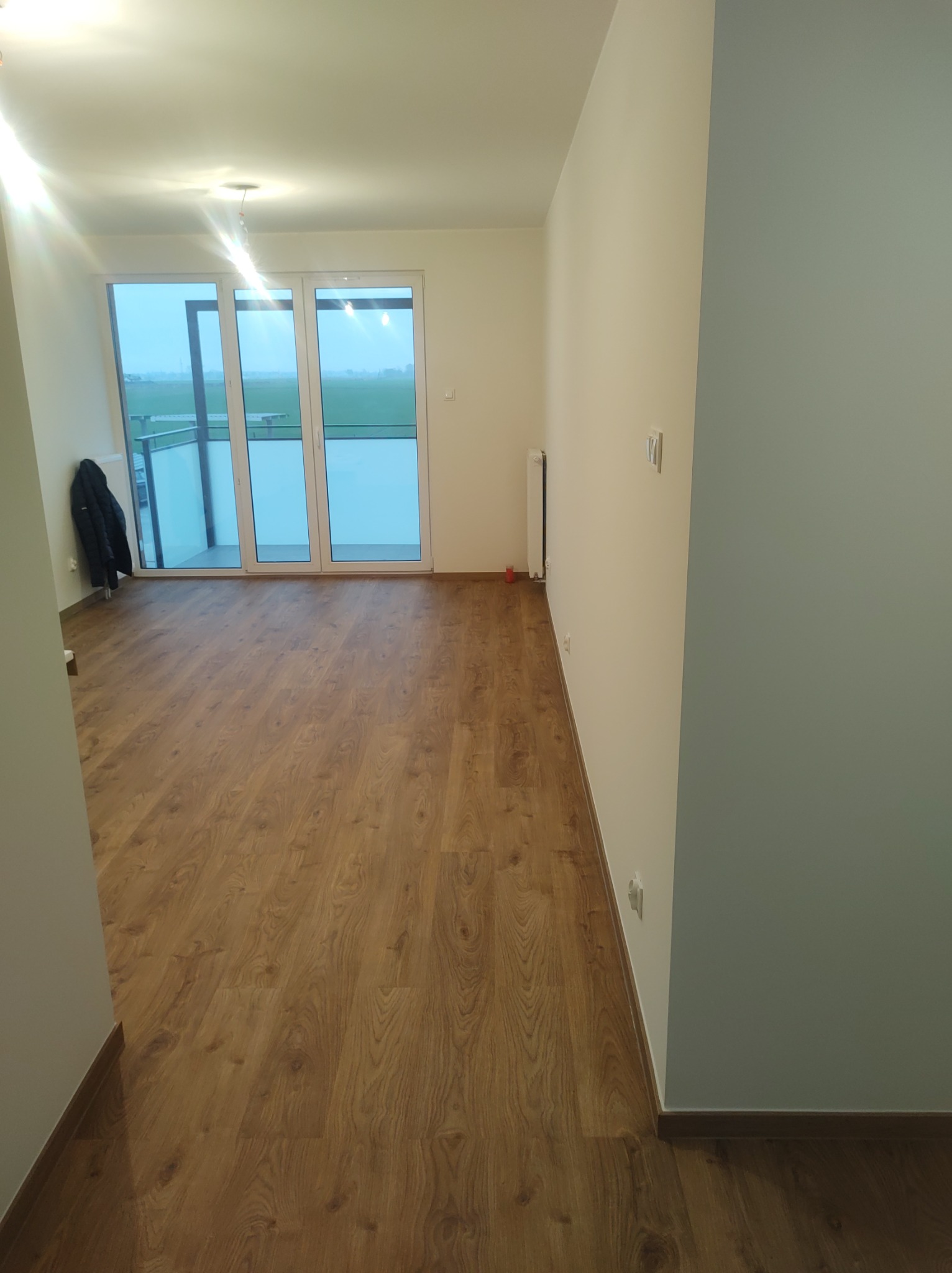 Jasne panele podłogowe w nowym mieszkaniu z balkonem, świeżo po remoncie. Widoczne gładkie, pomalowane ściany i grzejnik. Minimalistyczny design.